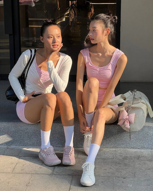 Two women in pink activewear and white sneakers sitting outside a fitness studio, sporty bag beside them
