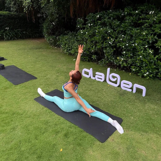 Woman in aqua activewear doing yoga splits outdoors on grass mat, lush greenery background