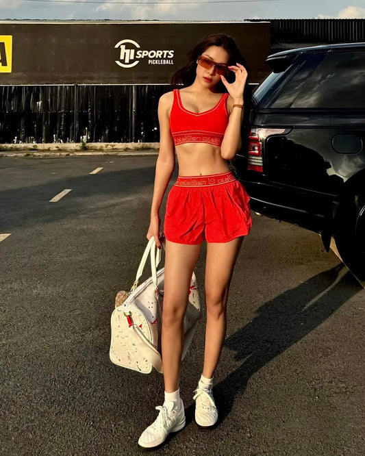 Woman in red athletic crop top and shorts with white sneakers, carrying a gym bag outdoors