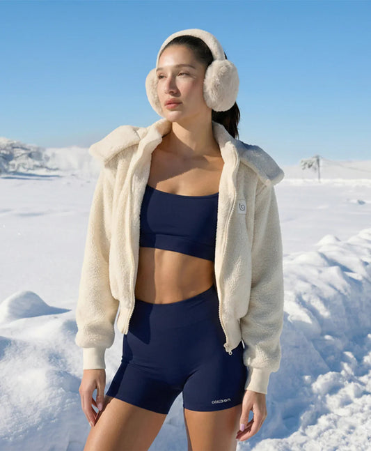 Woman in navy activewear and cream sherpa jacket with earmuffs standing in snowy landscape