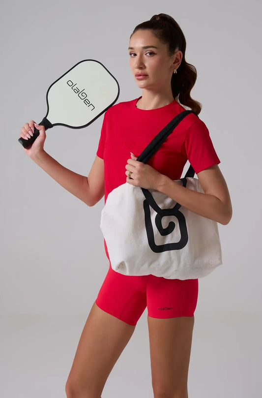 Woman in red embossed logo t-shirt and shorts holding paddle and white tote bag