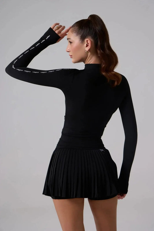Woman wearing black fitted long sleeve activewear top and pleated tennis skirt, rear view