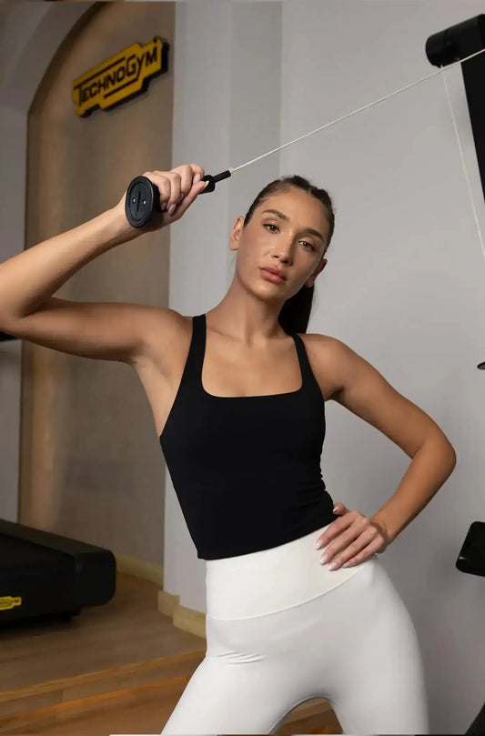 Woman wearing black racerback tank top and white leggings working out with cable machine in gym