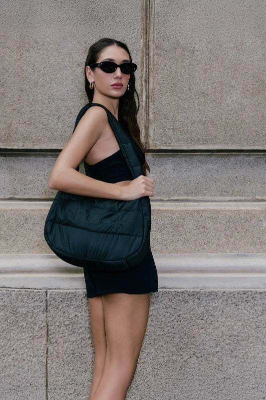Woman in black outfit with black padded tote bag, standing against stone wall