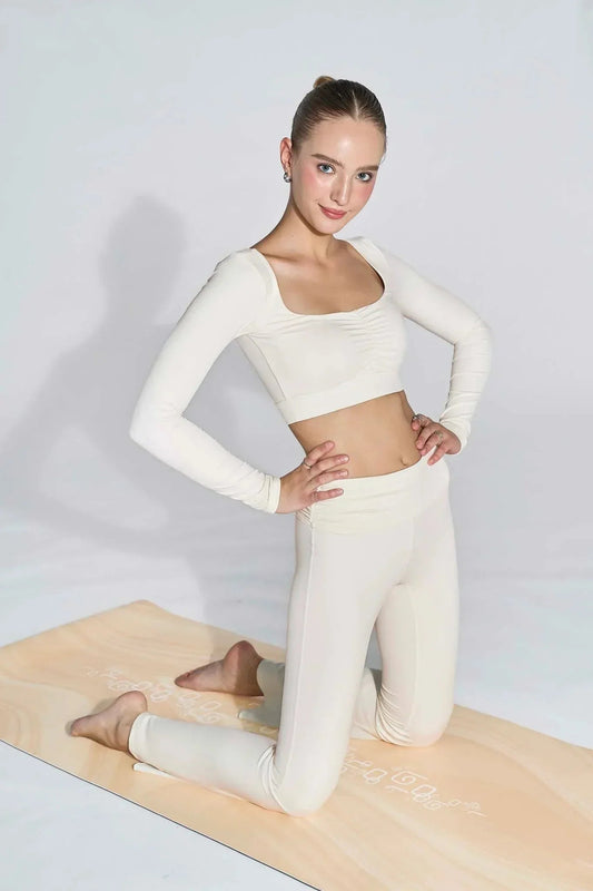 Woman in ivory white long sleeve ruched activewear top and leggings kneeling on a yoga mat