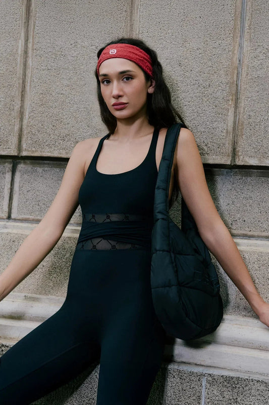 Woman in black monogram sleeveless jumpsuit with sheer mesh waistband, red headband, and black shoulder bag