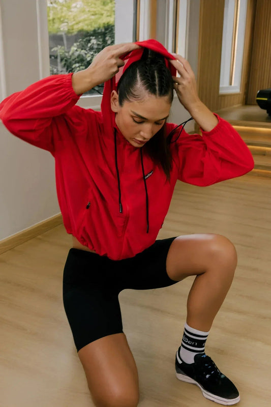 Woman in a red hooded workout jacket and black shorts kneeling in a gym setting